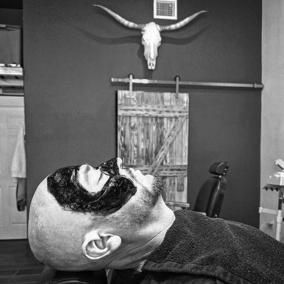 Man relaxing with a black facial mask in a barbershop, bull skull decor hanging on the wall.